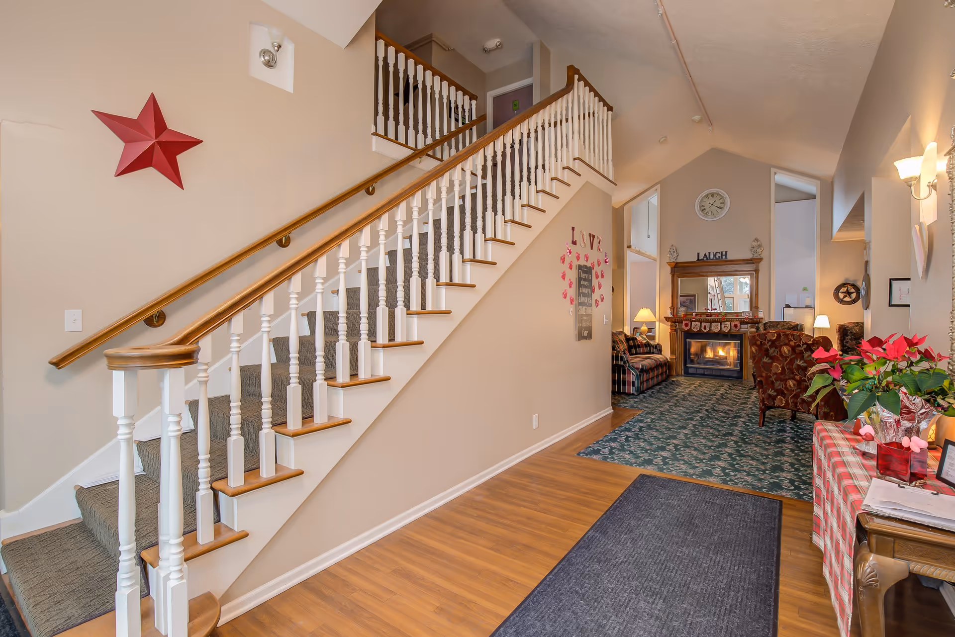 Interior view of a senior living facility showing a carpeted staircase with white railings and wooden handrails on the left. To the right, there is a hallway with wooden flooring leading to a cozy living room area with a fireplace, armchairs, a sofa, and a clock on the wall. The walls are decorated with a red star, a sign with the word LOVE and hearts, and other small decorations. A table with a red checkered cloth and a poinsettia plant is visible on the right side.
