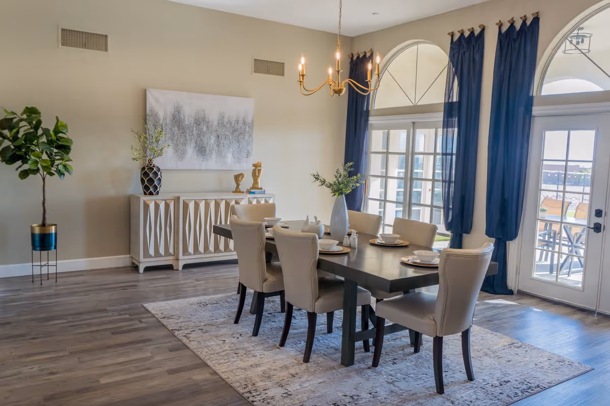 A modern dining room with a dark wooden table set for six with beige upholstered chairs. The room features large arched windows with dark blue curtains, a light fixture hanging above the table, a decorative sideboard with vases and sculptures, and a potted plant in the corner. The floor is wooden with a patterned area rug under the table.