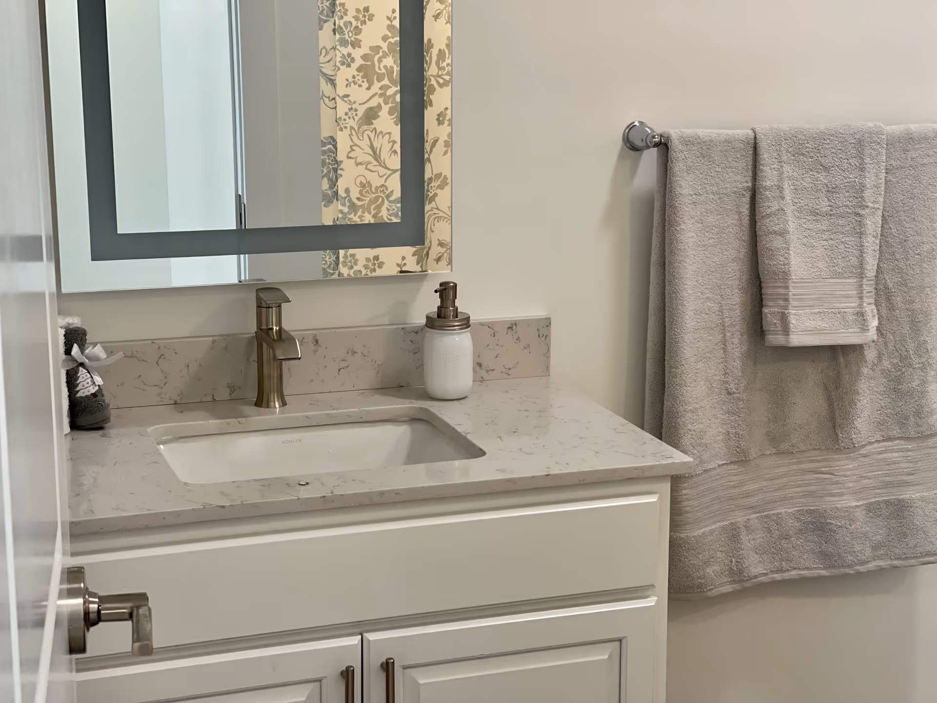 A clean and modern bathroom vanity with a white marble countertop, an undermount sink, a brushed nickel faucet, a soap dispenser, and a mirror above. To the right, a towel rack holds two beige towels, one folded over the other. The bathroom walls are painted white.
