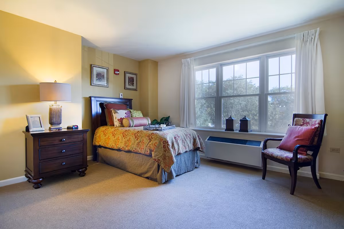 Well-lit furnished bedroom with a bed, nightstand and lamp, large window and an upholstered chair.