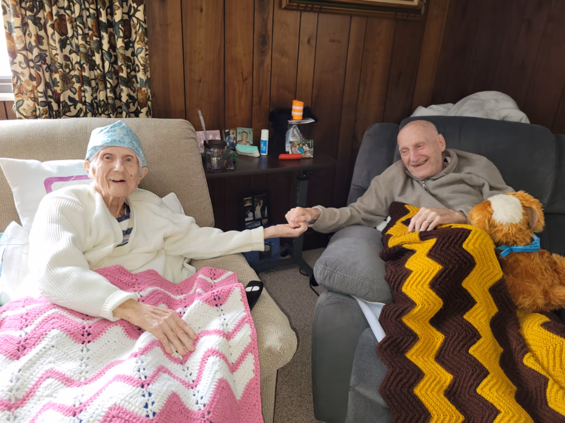 Two elderly men sitting in armchairs in a wood-paneled room, holding hands and smiling. One man is covered with a pink and white crocheted blanket and wearing a light blue cap, while the other has a brown and yellow crocheted blanket and a stuffed dog toy beside him.