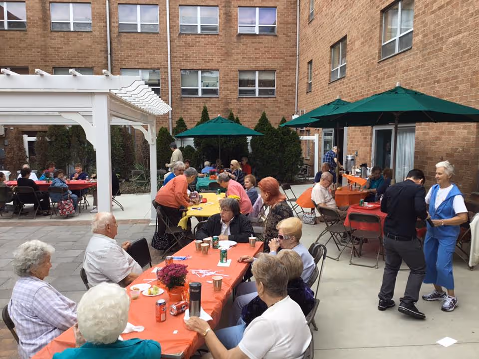 A group of elderly people sitting and socializing at tables covered with colorful tablecloths in an outdoor courtyard area of a senior living facility. There are green umbrellas providing shade, and some people are standing and interacting near the tables. The setting is surrounded by brick buildings and greenery.
