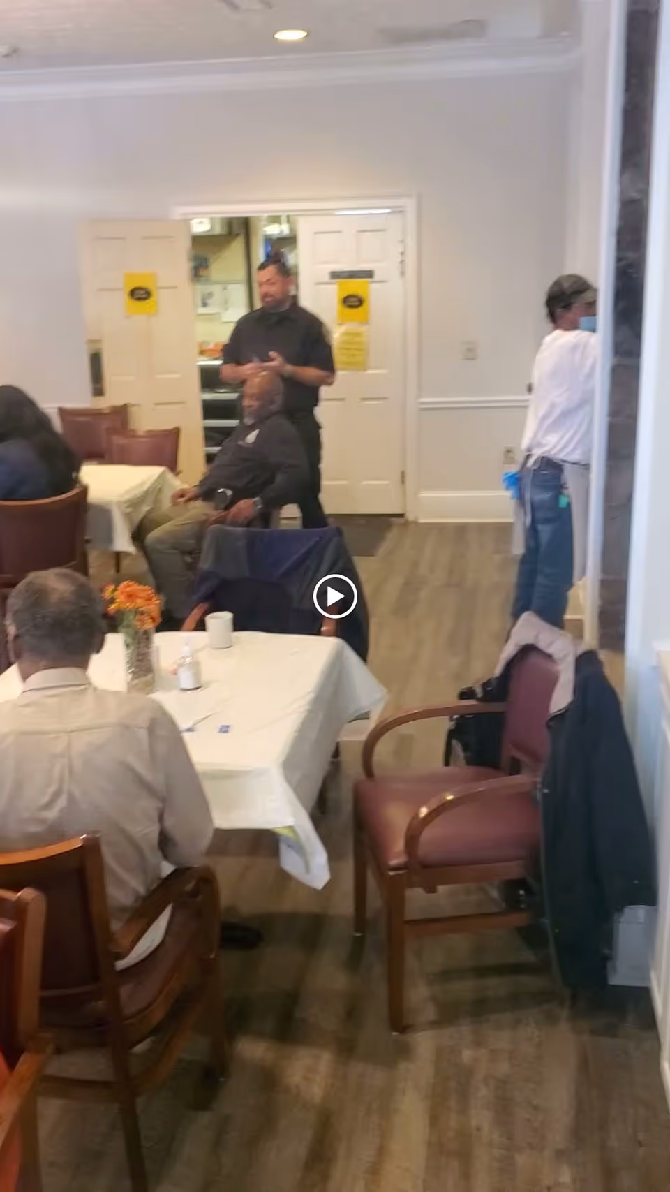 Communal dining room with tables, chairs, and several residents and staff near double doors.