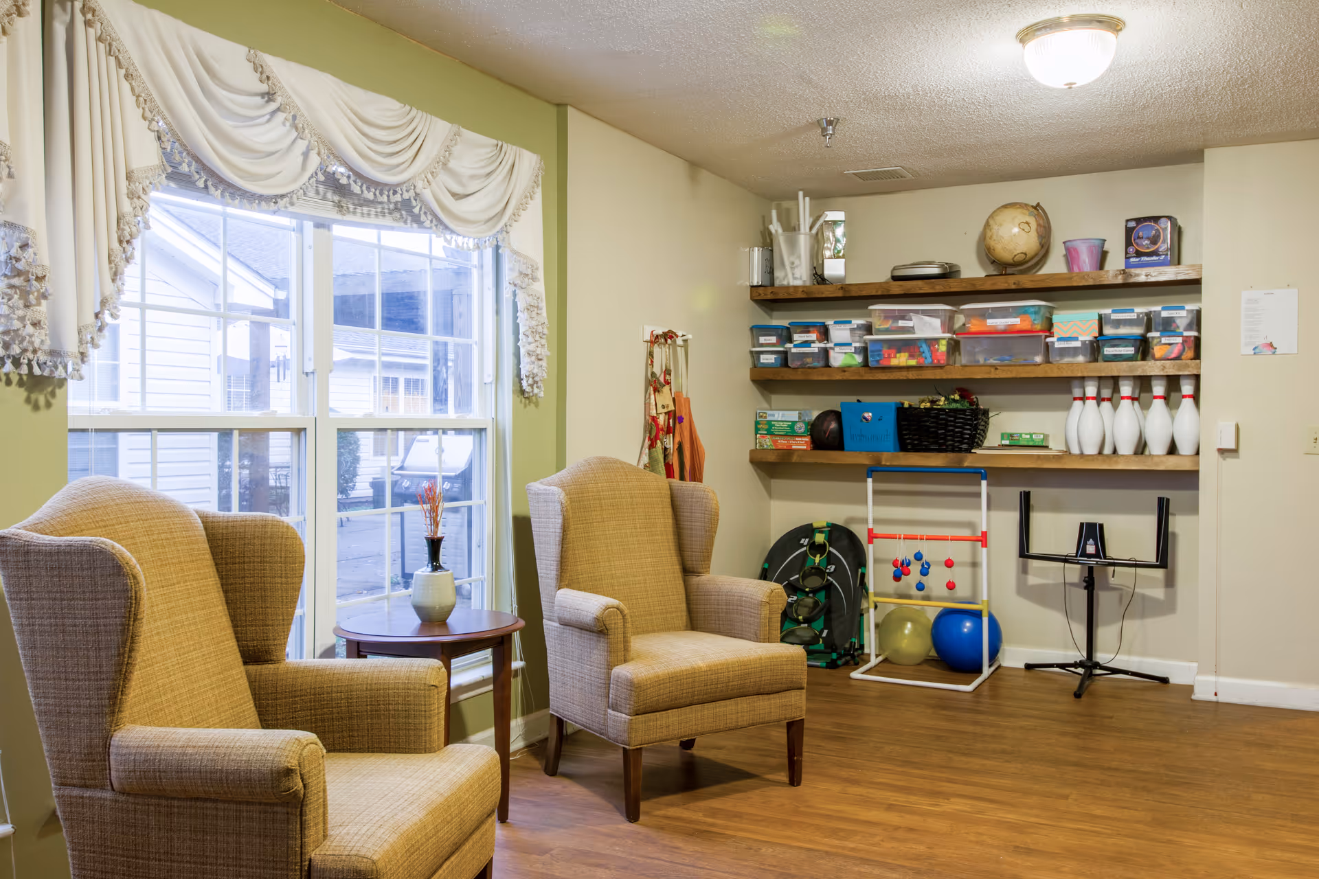 Cozy activity/living room with two upholstered armchairs by a large window and shelves stocked with games and bowling pins.