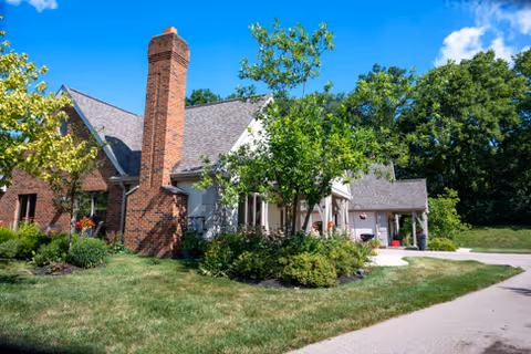 Front exterior of a brick and siding residential building with a prominent chimney, landscaped shrubs, driveway, and trees under a blue sky.