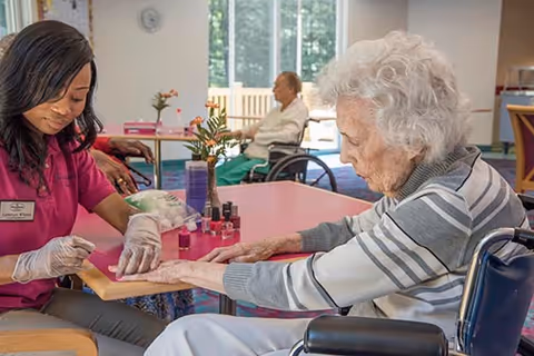 A caregiver wearing gloves is giving a manicure to an elderly woman in a wheelchair inside a bright room. Another elderly person in a wheelchair is visible in the background near large windows.