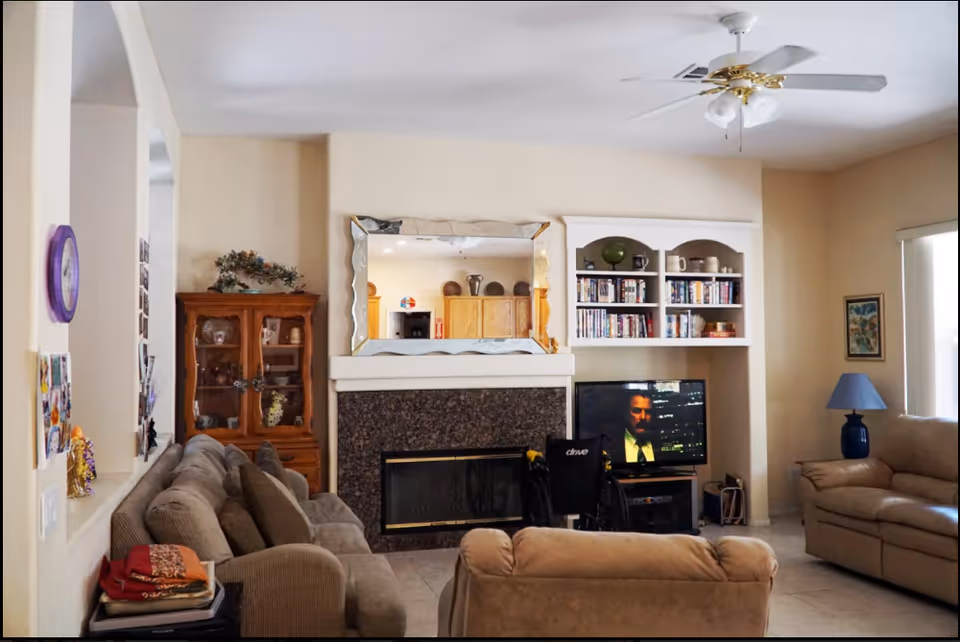 A cozy living room with beige walls and tile flooring. The room features a large beige sofa and a matching armchair, a fireplace with a decorative mirror above it, a TV on a stand showing a man in a suit, and a wheelchair positioned in front of the TV. There is a wooden cabinet with glass doors on the left side and built-in shelves with books and decorative items above the TV. A ceiling fan with lights is mounted on the ceiling, and a blue lamp sits on a side table next to another beige sofa near a window with vertical blinds.