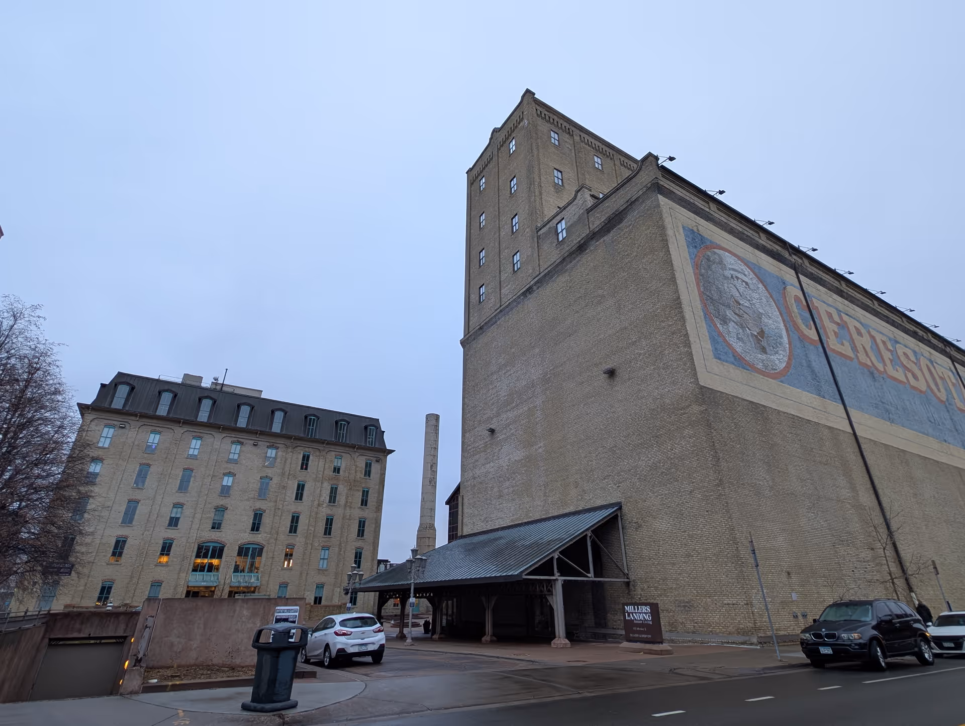 Exterior view of Millers Landing Senior Living facility showing a large multi-story brick building with a tall tower and a covered entrance. Several cars are parked along the street in front of the building under an overcast sky.
