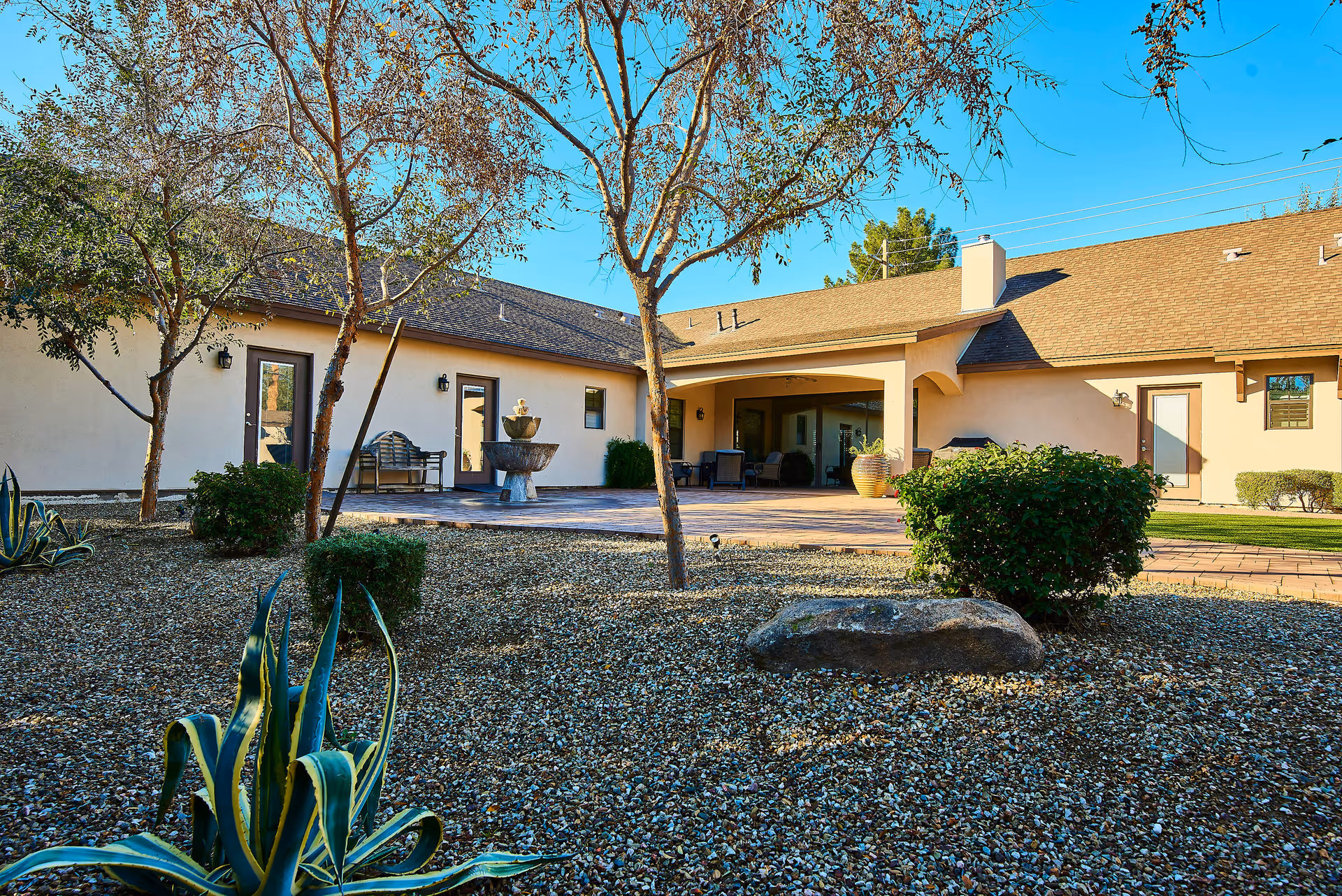 Outdoor view of a senior living facility courtyard with gravel ground, small bushes, agave plants, and leafless trees. The building has beige walls, multiple doors, and a covered patio area with seating and a water fountain.