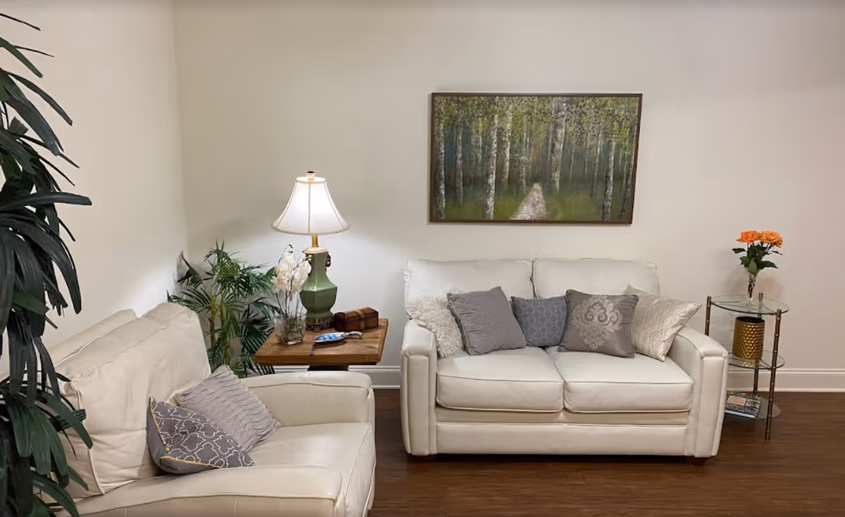 A cozy living room with two white leather sofas adorned with decorative pillows. A wooden side table with a green lamp, a small plant, and a decorative item is placed between the sofas. On the right, there is a glass side table with a vase of orange flowers and a golden decorative piece. A framed painting of a forest path hangs on the wall above the sofa. The room has wooden flooring and light-colored walls.