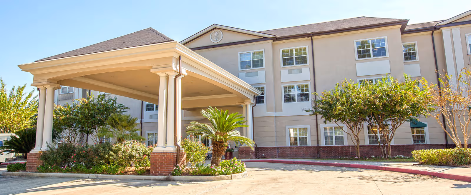 Exterior view of a multi-story senior living facility building with a covered entrance supported by columns, surrounded by landscaped greenery and trees under a clear blue sky.