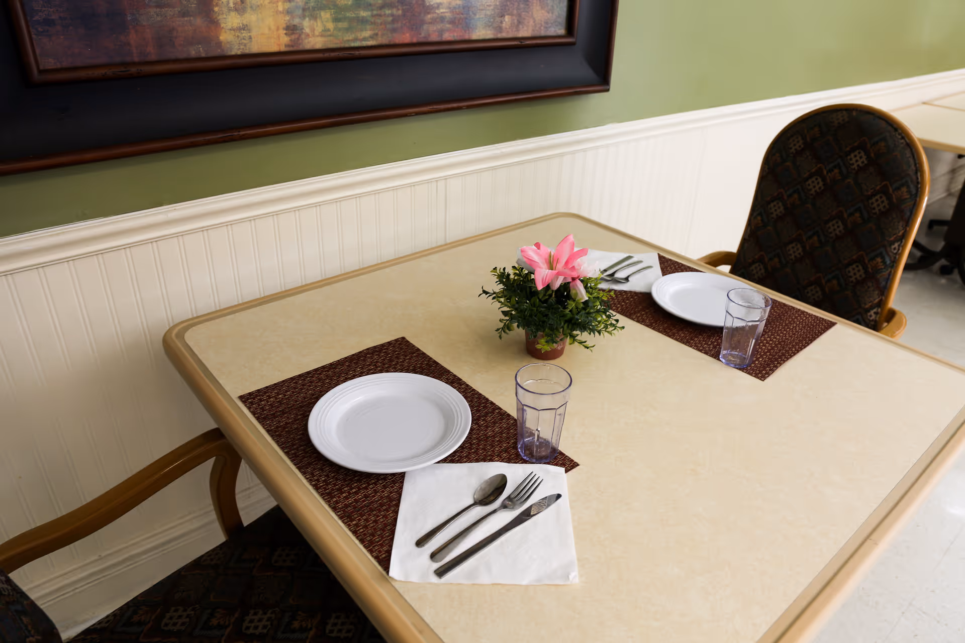 A small dining table set for two with plates, utensils, plastic cups and a potted flower between two upholstered chairs against a beadboard wall.