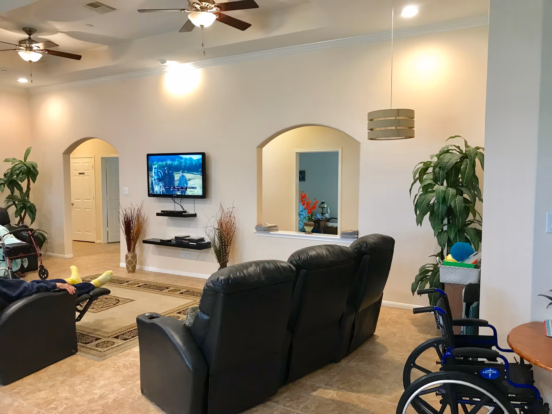 A living room area in an assisted living facility with three black recliner chairs facing a wall-mounted TV. The room has beige walls, tile flooring, ceiling fans with lights, and decorative plants. A wheelchair is visible near a small round table on the right side. There are two arched openings in the wall, one showing a hallway and the other a room with a mirror and flowers.