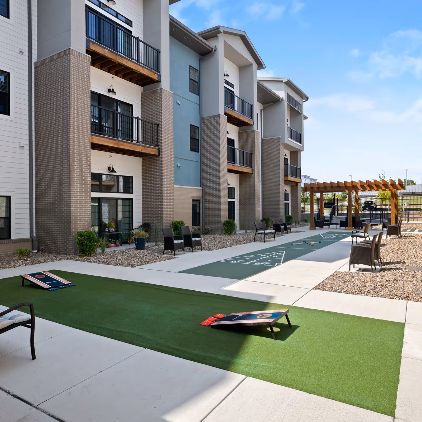 Outdoor recreational area at a senior living facility featuring a shuffleboard court, cornhole boards, chairs, and a pergola with seating, adjacent to a multi-story building with balconies and large windows under a clear blue sky.