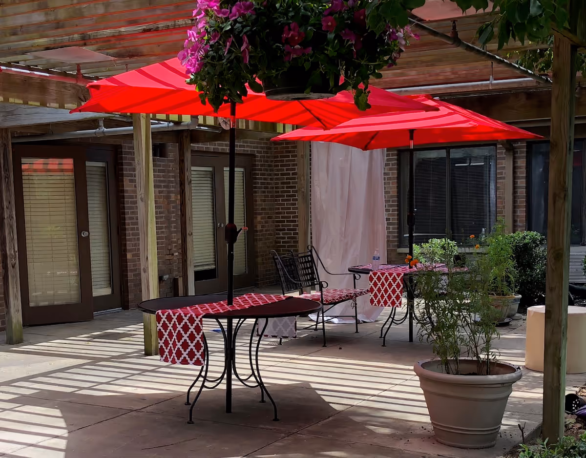 Outdoor patio area with red umbrellas over tables covered with red and white patterned table runners. There are metal chairs, potted plants, and hanging flower baskets. The area is shaded by a wooden pergola and surrounded by brick walls with windows and doors.