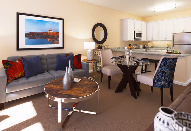 A bright and cozy living area in a senior living facility featuring a gray sofa with colorful pillows, a round wooden coffee table with decorative vases, a small dining table set with four chairs, and a kitchen area with white cabinets, a microwave, and a stainless steel refrigerator. The room is warmly lit with a table lamp and natural light coming through a window.