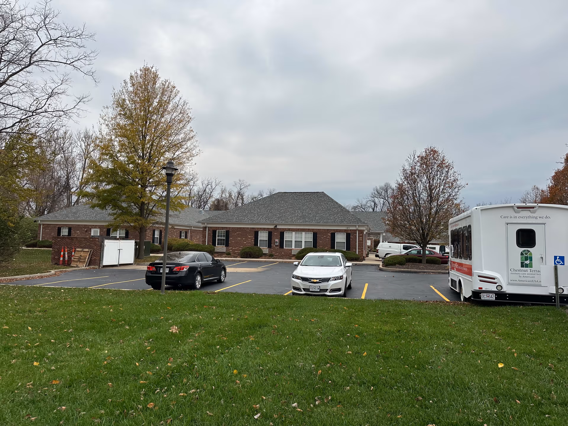 Brick senior living building with a parking lot containing cars and a shuttle bus under an overcast sky.