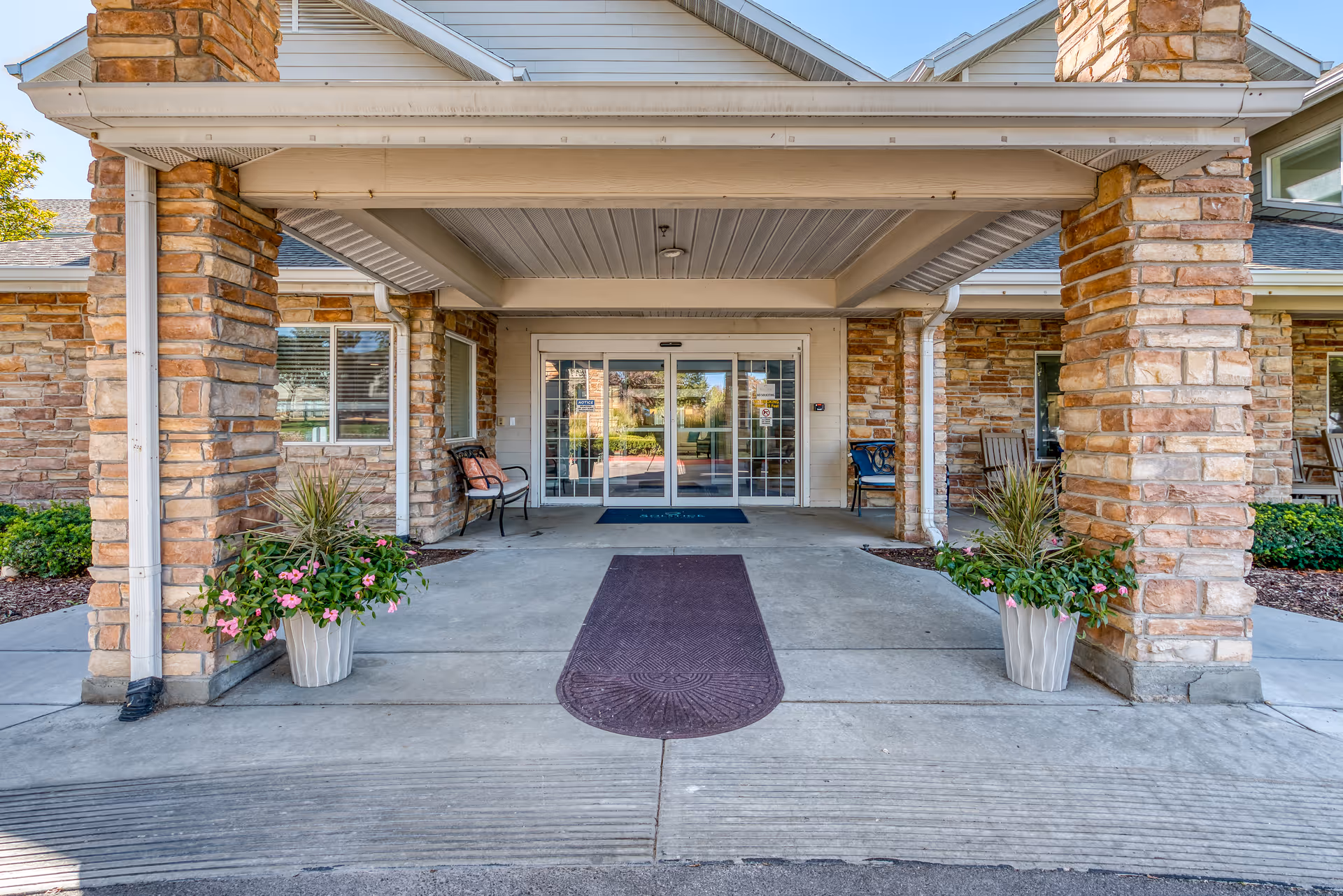 Entrance of a senior living facility with stone pillars, a covered walkway, potted plants with pink flowers, benches on either side, and glass double doors leading inside.