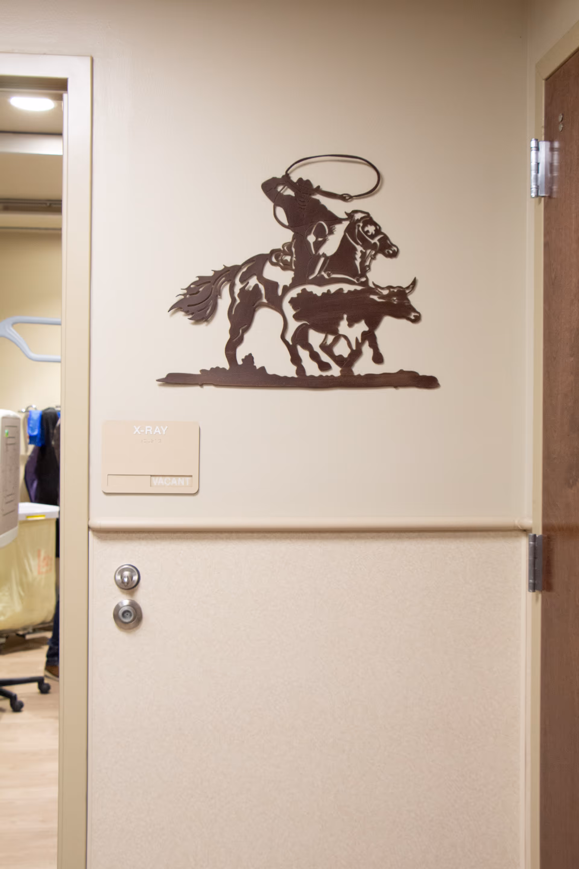 Interior hallway wall with a metal art piece depicting a cowboy on a horse roping a calf. To the left is an open door leading to an X-ray room, indicated by a sign that reads 'X-RAY' with a smaller sign below it stating 'VACANT'.