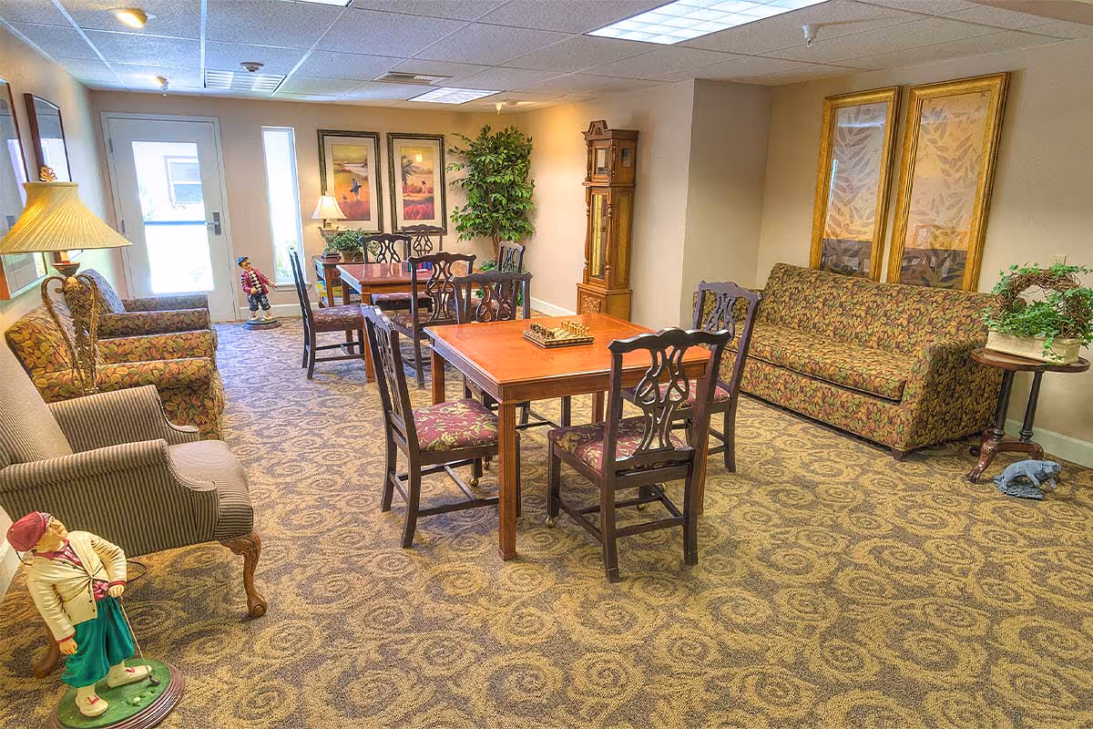A cozy common area in a senior living facility featuring patterned carpet, a wooden table with four chairs, a floral-patterned sofa, armchairs, a grandfather clock, framed artwork on the walls, potted plants, and decorative figurines. The room is well-lit with ceiling lights and lamps, and there is a door with windows letting in natural light.