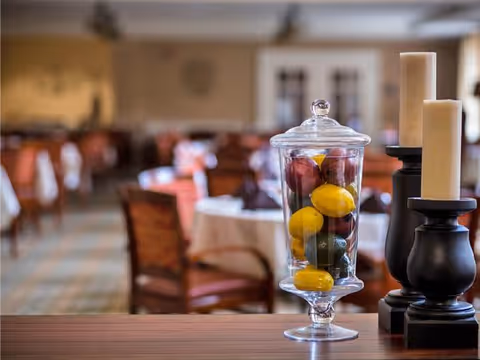 Glass apothecary jar filled with decorative fruit and pillar candles on a table in a softly focused dining room.