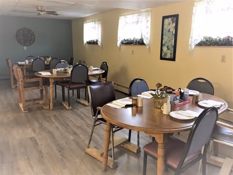 A dining room with several wooden tables and chairs arranged for meals. Each table has place settings with plates, napkins, and utensils. The room has light yellow walls with three windows adorned with floral curtains and small flower pots on the windowsills. There is a framed floral picture on the wall and a decorative round wall piece in the background.