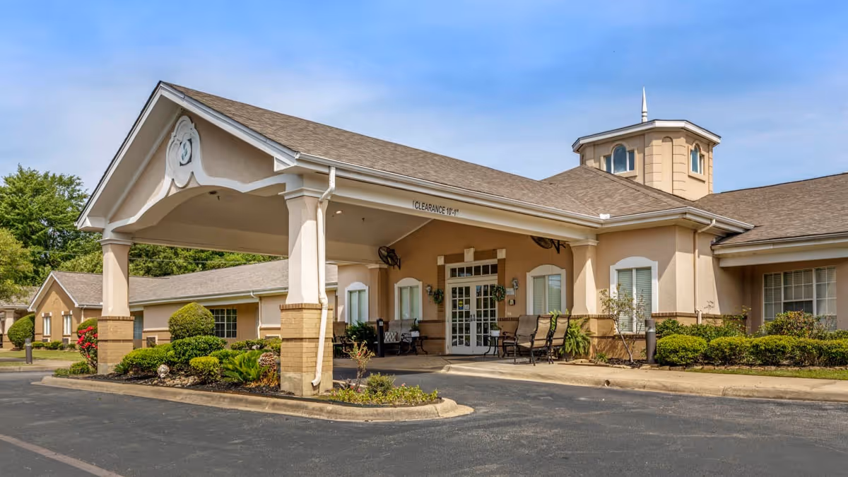 Exterior view of Brookdale Henderson senior living facility showing the main entrance with a covered drop-off area, beige walls, and a small tower feature on the roof. There are shrubs and landscaped plants around the building and a paved driveway in front.