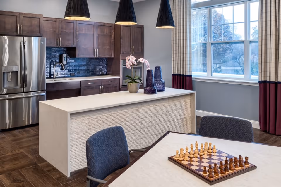 Modern kitchen and dining area with a central island, stainless refrigerator, pendant lights, and a chessboard on a table.
