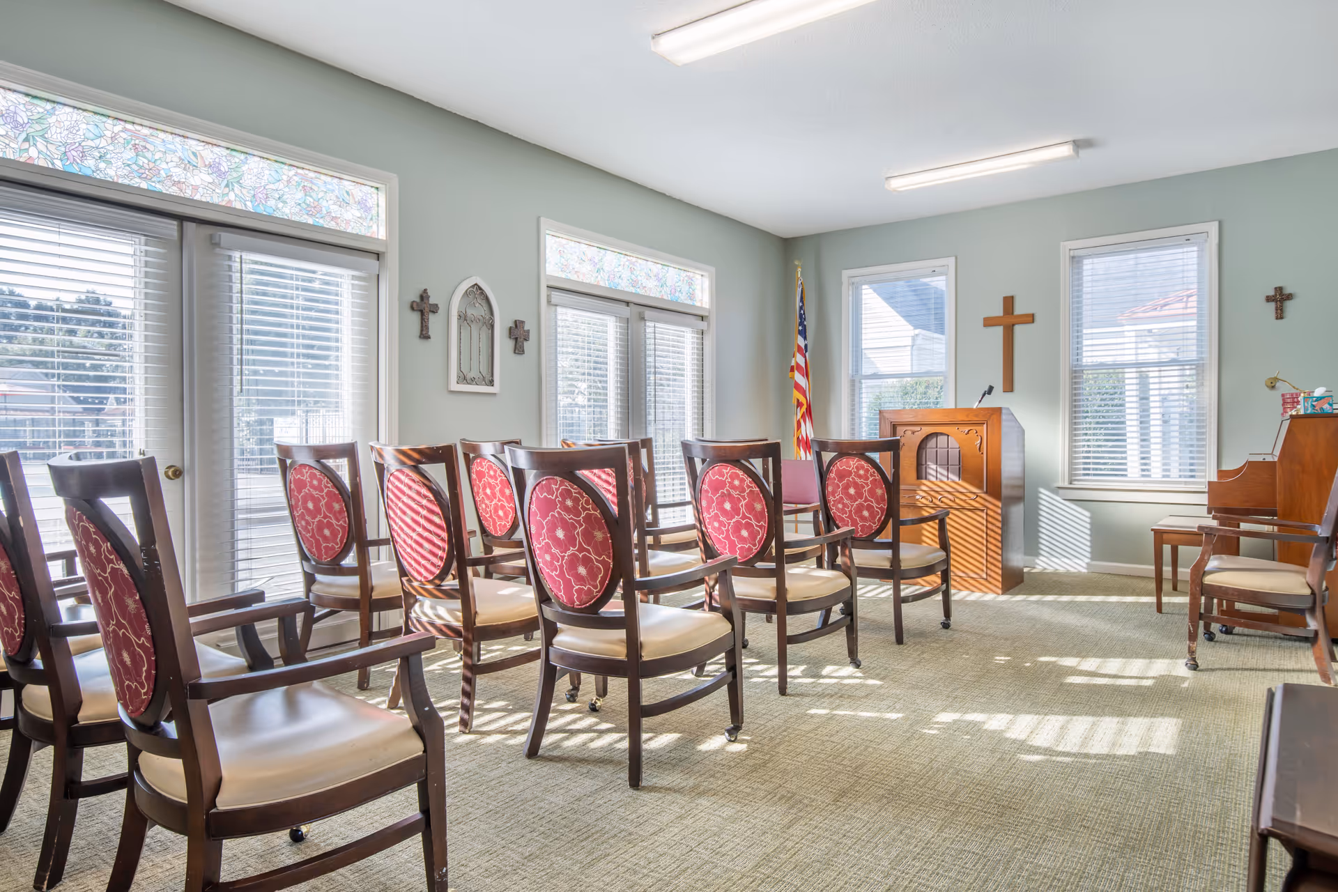A small chapel or meeting room with rows of wooden chairs featuring red patterned cushions. The room has light green walls, several windows with white blinds, and stained glass panels above the doors. There is a wooden podium with a microphone, an American flag, a piano, and multiple crosses on the walls.