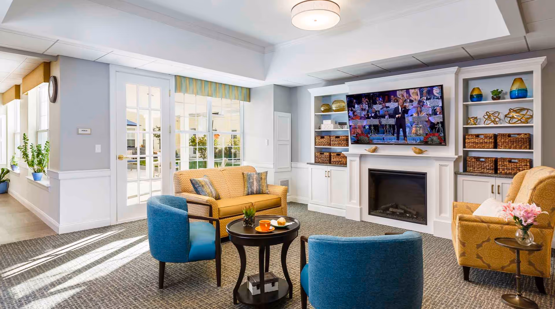 A bright and cozy living room in a senior living facility with a yellow sofa, two blue armchairs, and a yellow armchair arranged around a small round black coffee table. A flat-screen TV is mounted above a white fireplace with built-in shelves on either side, decorated with baskets, vases, and decorative objects. Large windows and glass doors allow natural light to fill the room, and a small plant is placed on a side table next to the yellow armchair.