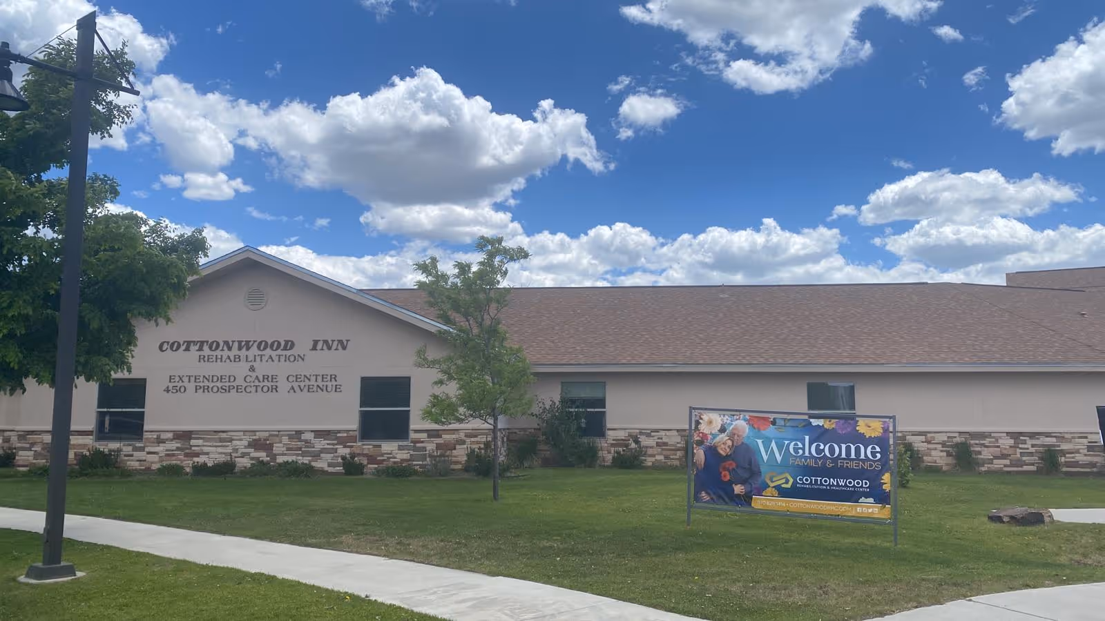 Exterior view of Cottonwood Inn Rehab and Extended Care Center building with a sign on the lawn that reads 'Welcome Family & Friends Cottonwood'. The building has a beige facade with stone accents along the bottom and a brown roof. The sky is blue with scattered white clouds.