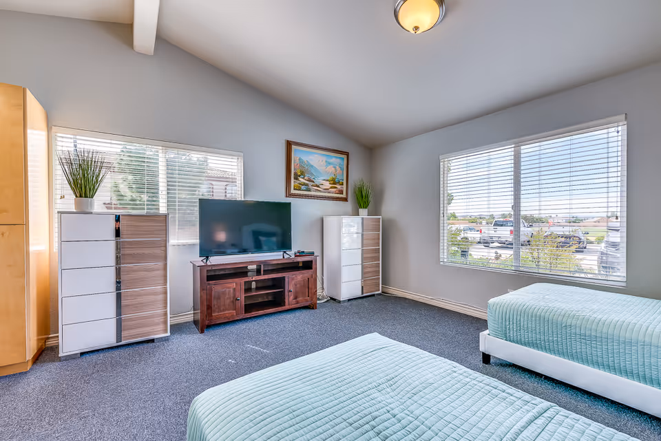 Bright bedroom with two twin beds, a TV on a wooden stand, dressers, and large windows letting in daylight.