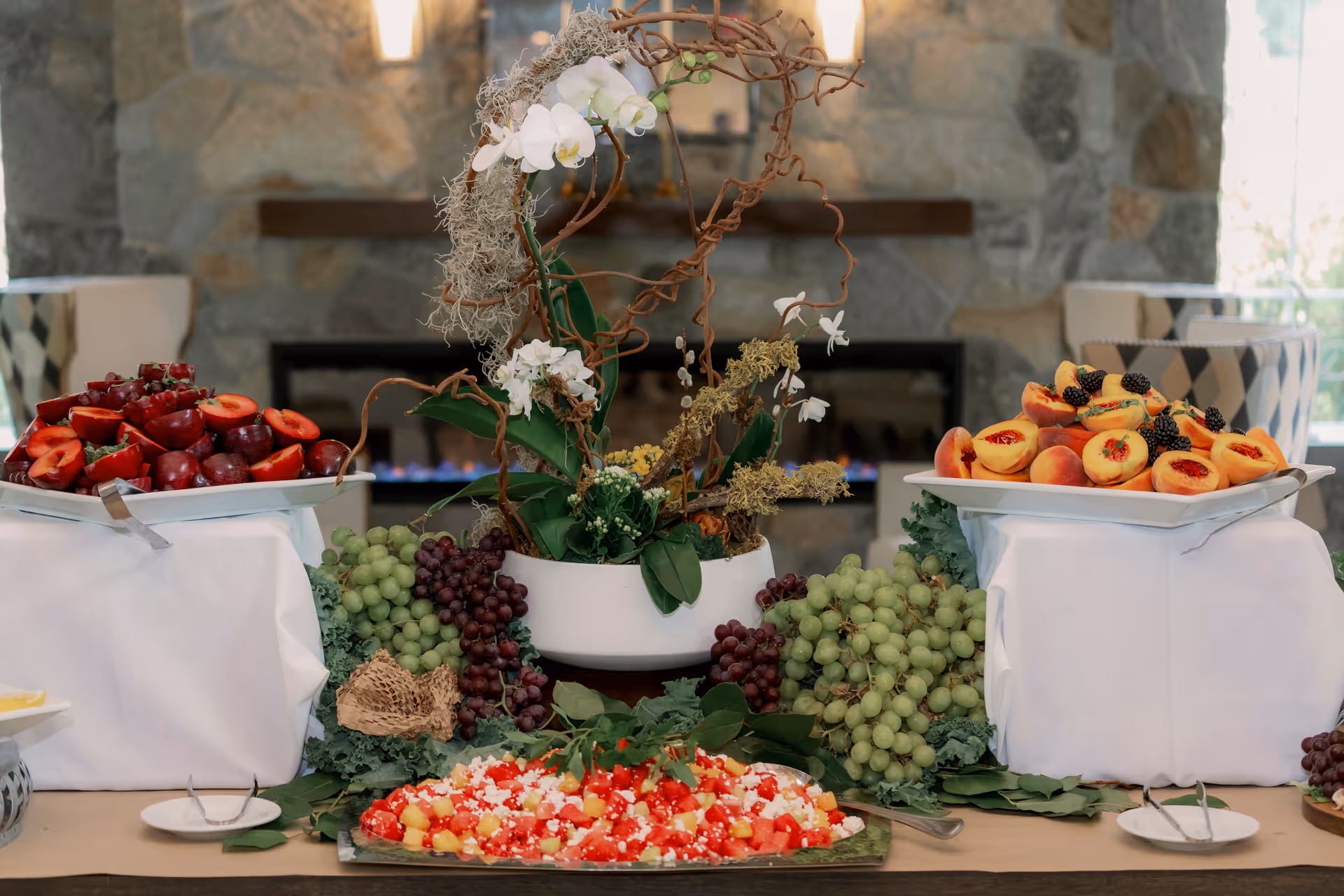 A buffet table with an assortment of fresh fruits including grapes, plums, and halved peaches topped with blackberries, arranged around a decorative floral centerpiece in a room with a stone fireplace and cushioned chairs in the background.