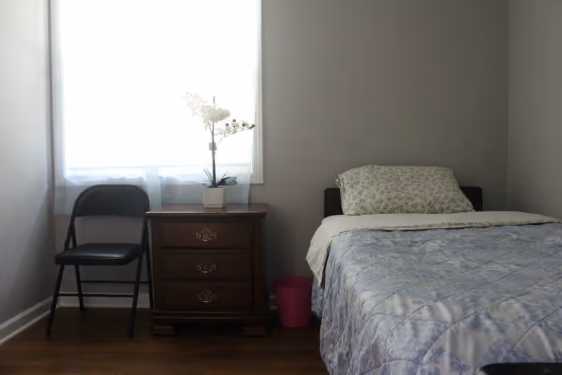 A simple bedroom with a single bed covered in a blue patterned quilt and a floral pillow. Next to the bed is a wooden nightstand with three drawers, topped with a small potted white orchid. A black folding chair is placed beside the nightstand in front of a window with sheer white curtains. The walls are painted light gray and the floor is wooden.