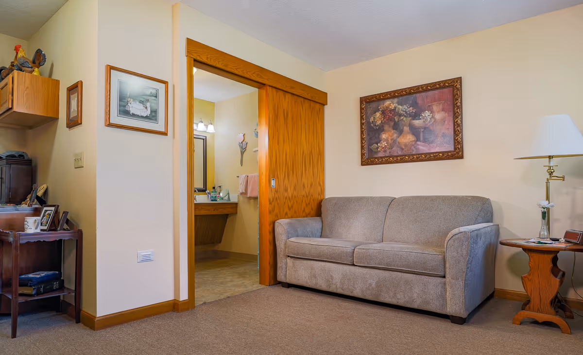 A cozy living room area with a beige couch, a wooden side table with a lamp and a small flower vase, and a framed painting of flowers on the wall. To the left, there is an open sliding wooden door leading to a bathroom with a vanity mirror and sink. The room has beige walls and carpeted floor, with some decorative items and framed pictures on the left side.