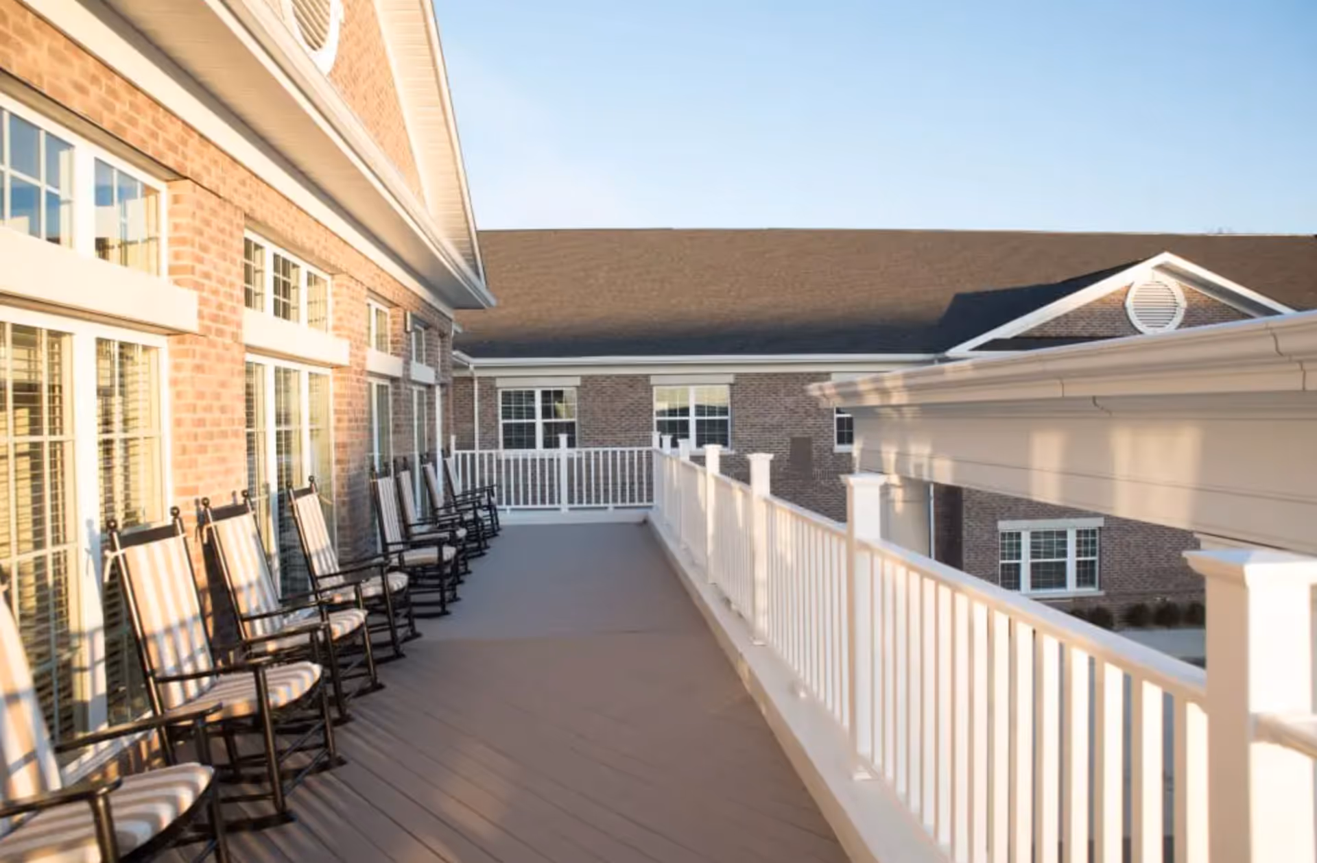 A sunlit outdoor balcony with a row of striped cushioned rocking chairs along a white railing beside a brick building.