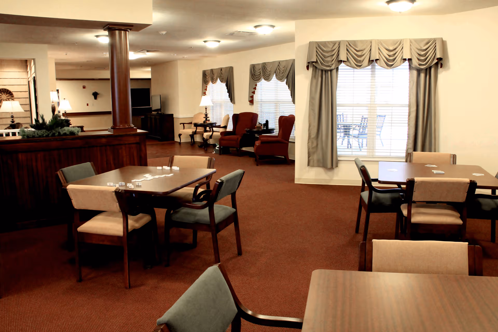 Interior of a senior living facility common area with multiple tables and chairs arranged for games or socializing. There are two red armchairs near windows with draped curtains, and a television is visible in the background. The room has warm lighting and carpeted floors.