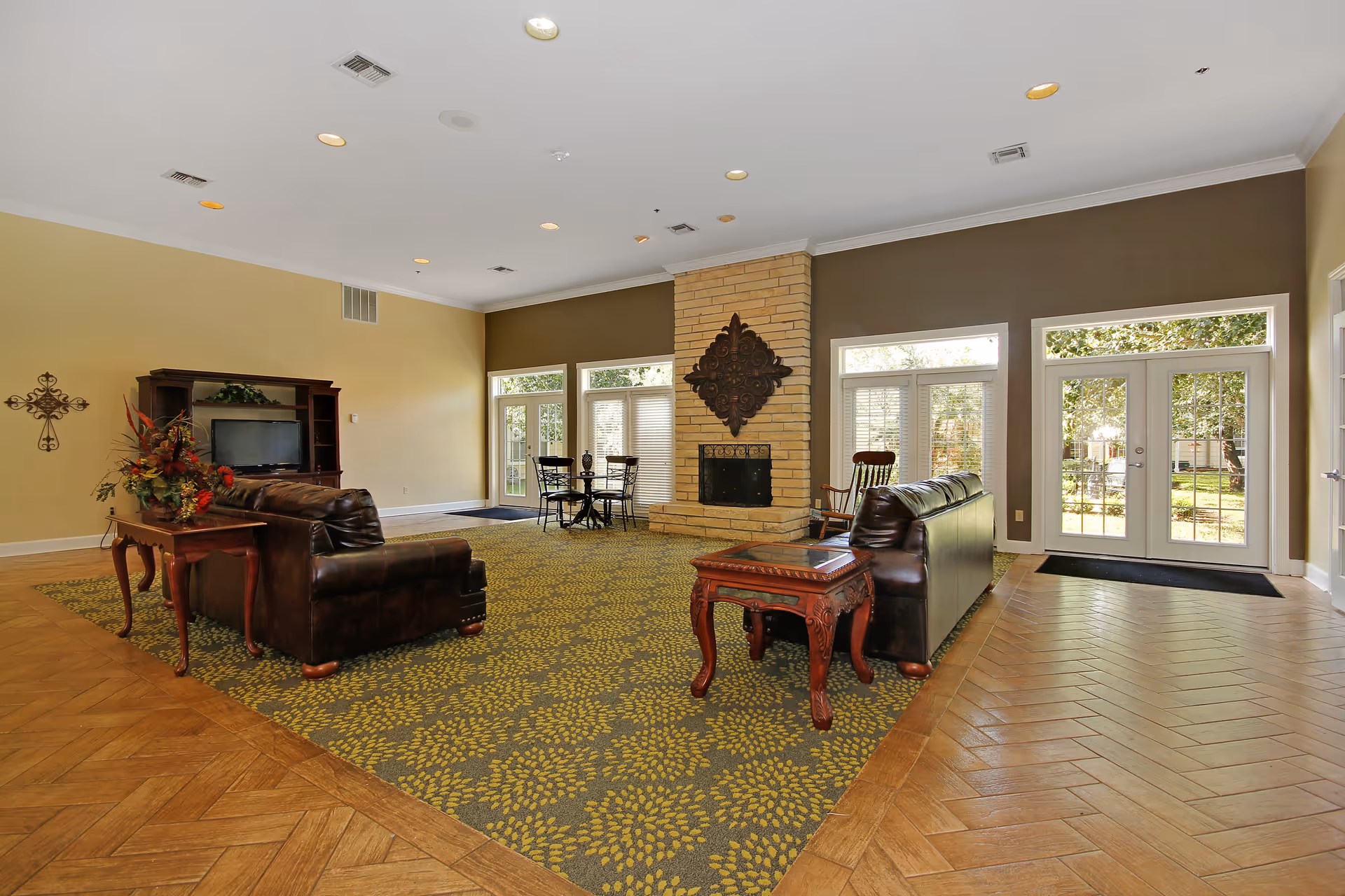 A spacious living room area with two dark brown leather sofas facing each other, a wooden side table with a floral arrangement, a TV on a wooden entertainment center, a stone fireplace with decorative metal art above it, and large windows and glass doors letting in natural light and showing greenery outside.