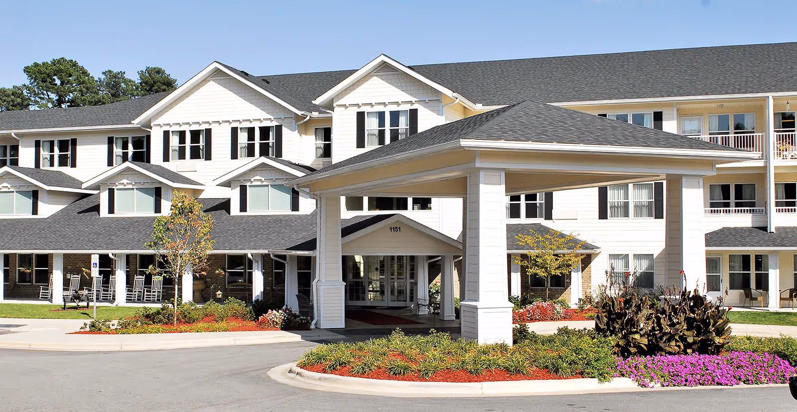 Front exterior view of The Lodge at Wake Forest Gracious Retirement Living, showing a large multi-story building with white siding, black shutters, and a covered entrance. There are landscaped flower beds with green shrubs and purple flowers in front of the entrance, and a paved driveway leading up to the building.
