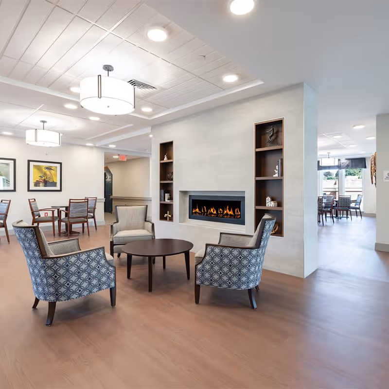 A modern senior living community common area with a cozy fireplace set into a wall with built-in shelves. Four upholstered armchairs with patterned fabric surround a round wooden coffee table. In the background, there are dining tables and chairs, framed artwork on the walls, and large windows letting in natural light.