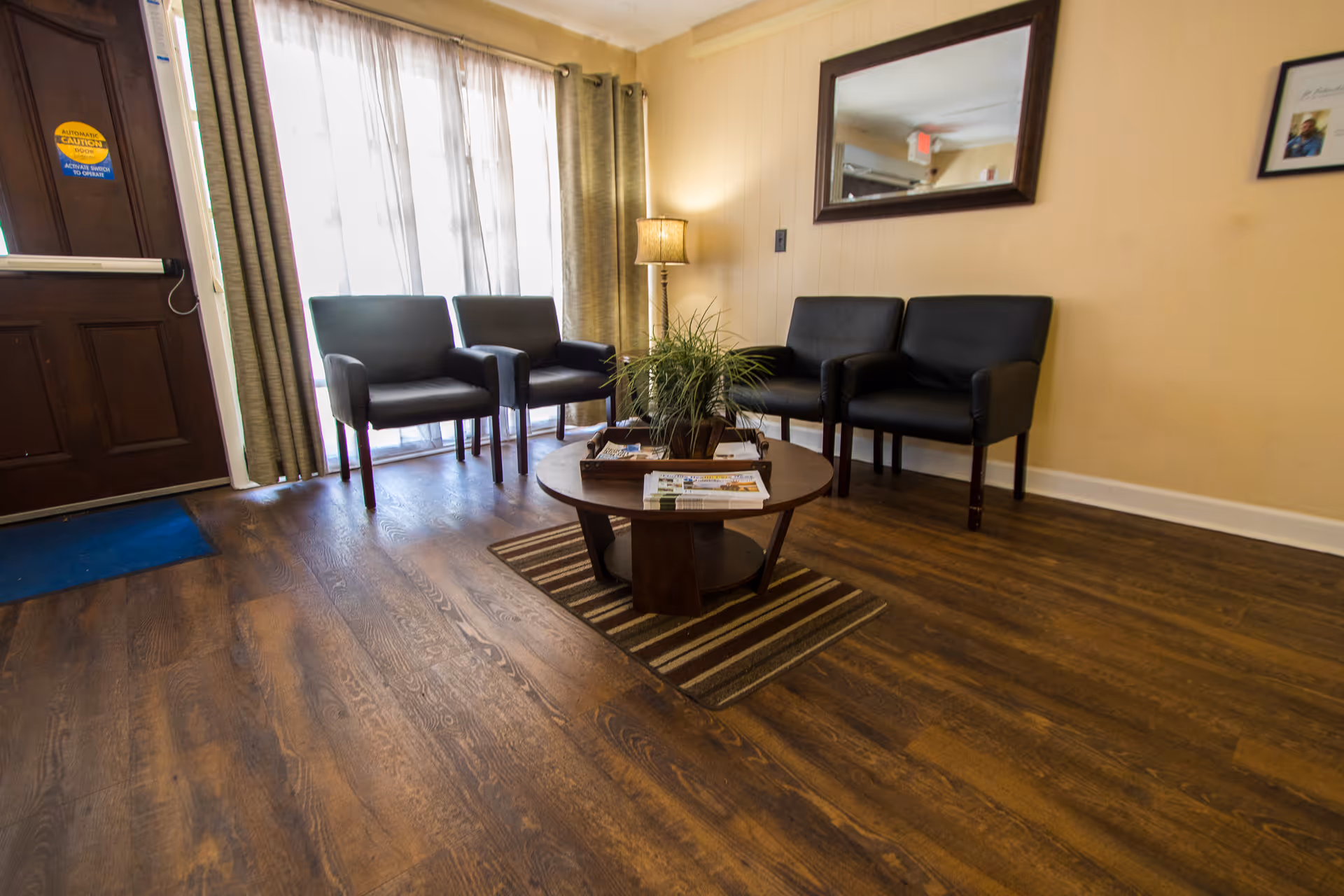 Small nursing facility waiting room with black chairs, a round coffee table holding magazines and a potted plant, a mirror on the wall, and wood floors.
