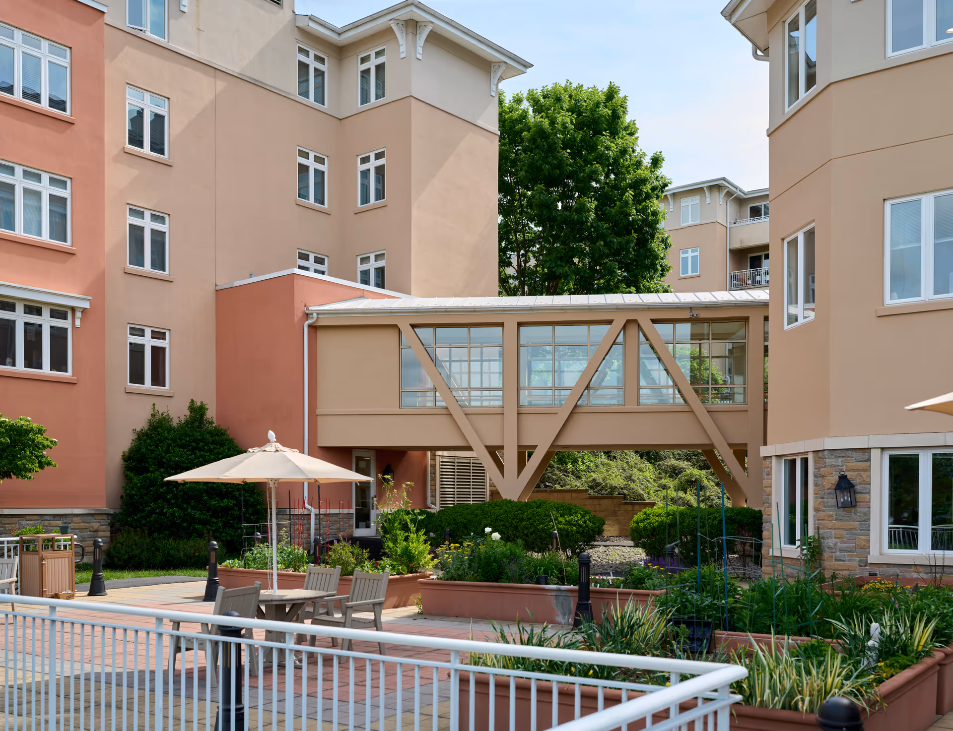 Outdoor courtyard area of a senior living facility with beige and peach-colored buildings surrounding a garden with green plants and flowers. There is a covered walkway bridge connecting two buildings, patio tables with umbrellas, and a white railing in the foreground.