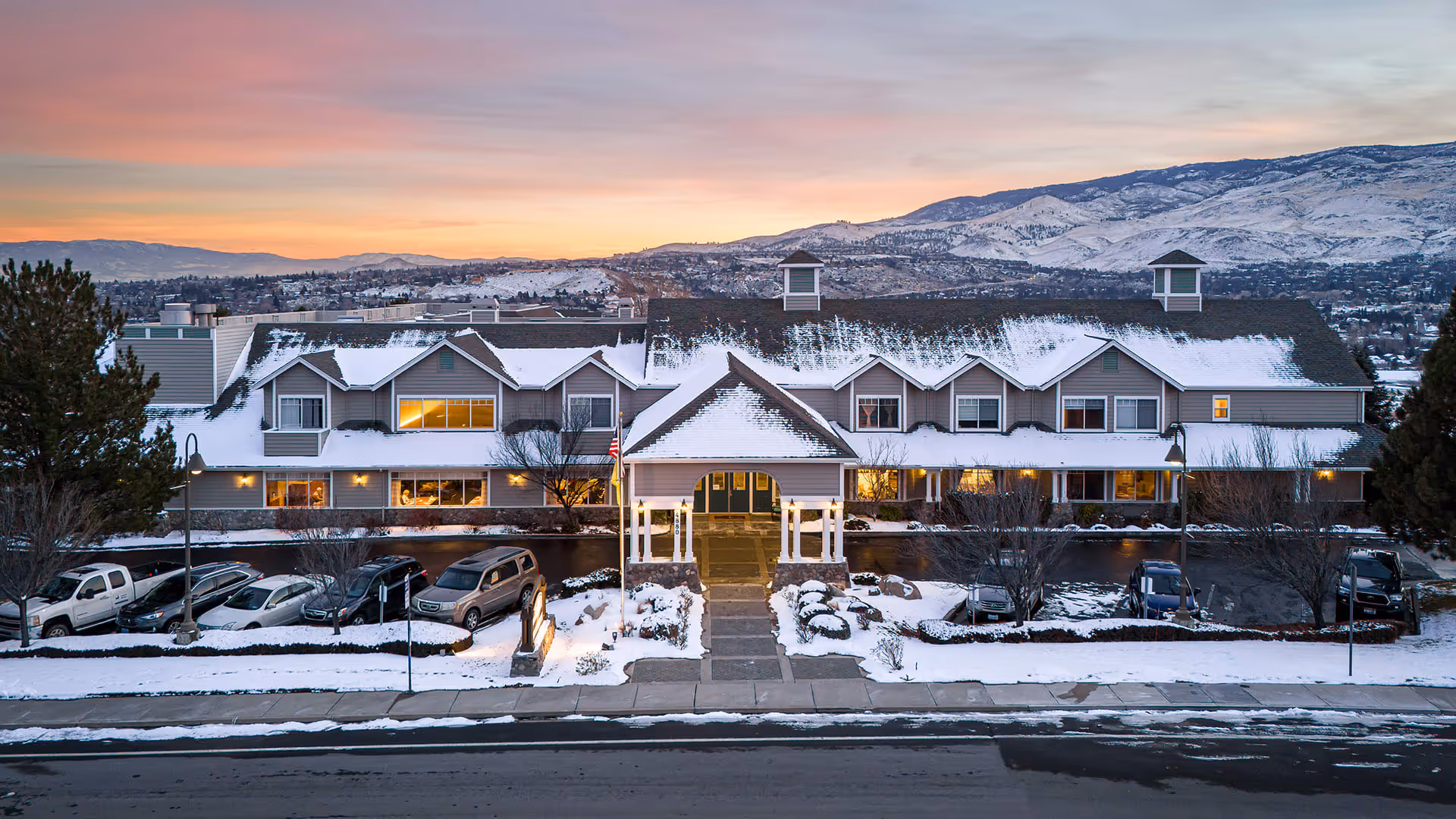 Exterior view of a senior living facility named Atria Summit Ridge during winter at sunset, with snow on the roof and ground, several parked cars in front, and mountains in the background.
