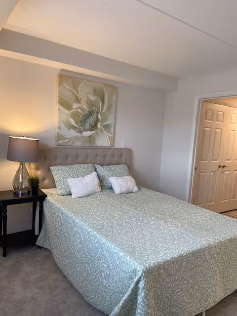 A bedroom with a neatly made bed covered in a light green patterned bedspread and matching pillows. There is a beige tufted headboard, a nightstand with a lamp and a small plant, and a large floral painting hanging on the wall above the bed. A doorway with double doors is visible on the right side of the room.