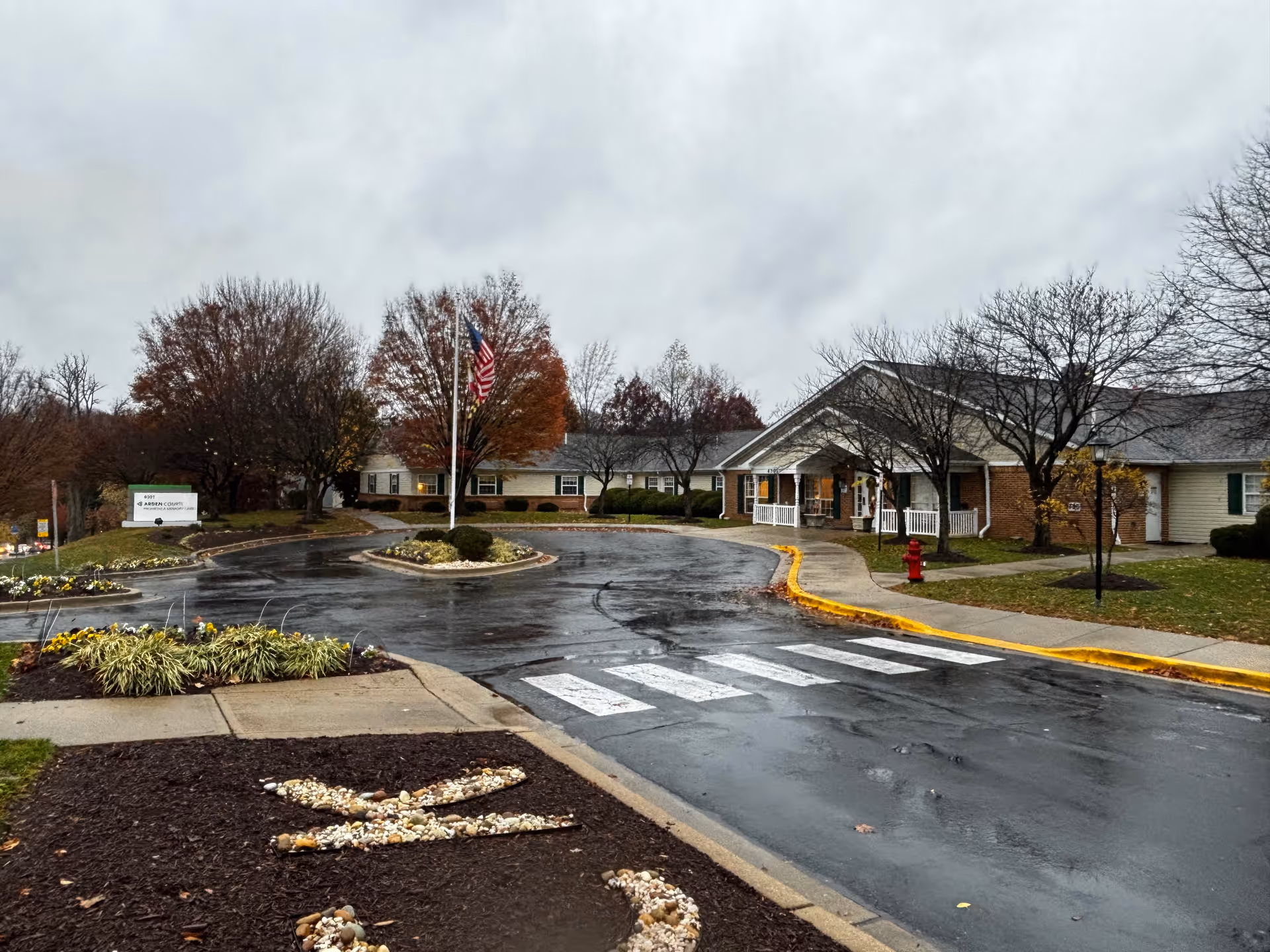 Exterior view of Arden Courts - ProMedica Memory Care Community (Kensington) on a cloudy day. The building is a single-story structure with a covered entrance and white railings. There is a circular driveway with a crosswalk and a flagpole with an American flag in the center. Trees with autumn foliage surround the area, and there are landscaped flower beds near the driveway.