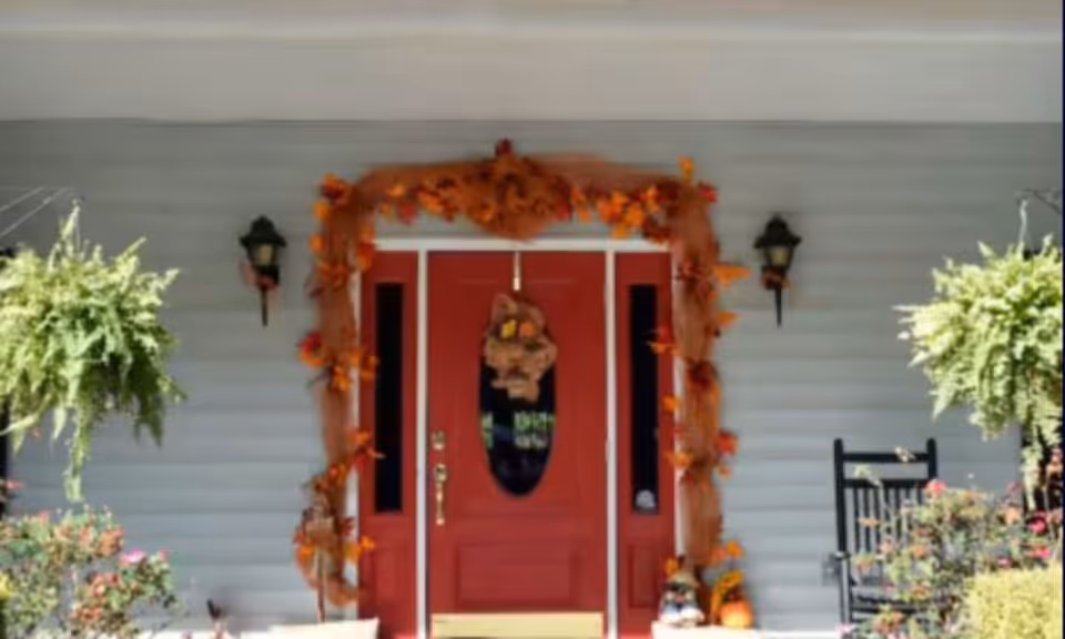 Front entrance of a building with a red door decorated with autumn-themed garlands and a wreath. Two black wall-mounted lanterns flank the door. There are green hanging plants on either side and a black rocking chair on the right side of the porch.