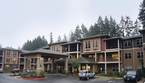 Front exterior of a three-story senior living building with a covered porte-cochere, parked cars, and tall trees in the background.