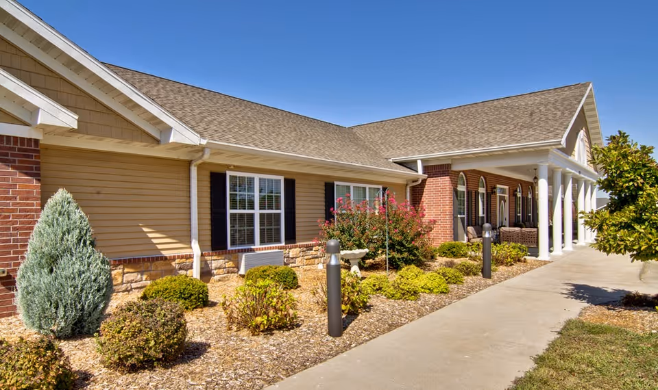 Exterior view of a senior living facility building with a beige and brick facade, white columns along a covered porch, landscaped bushes and plants, and a clear blue sky.