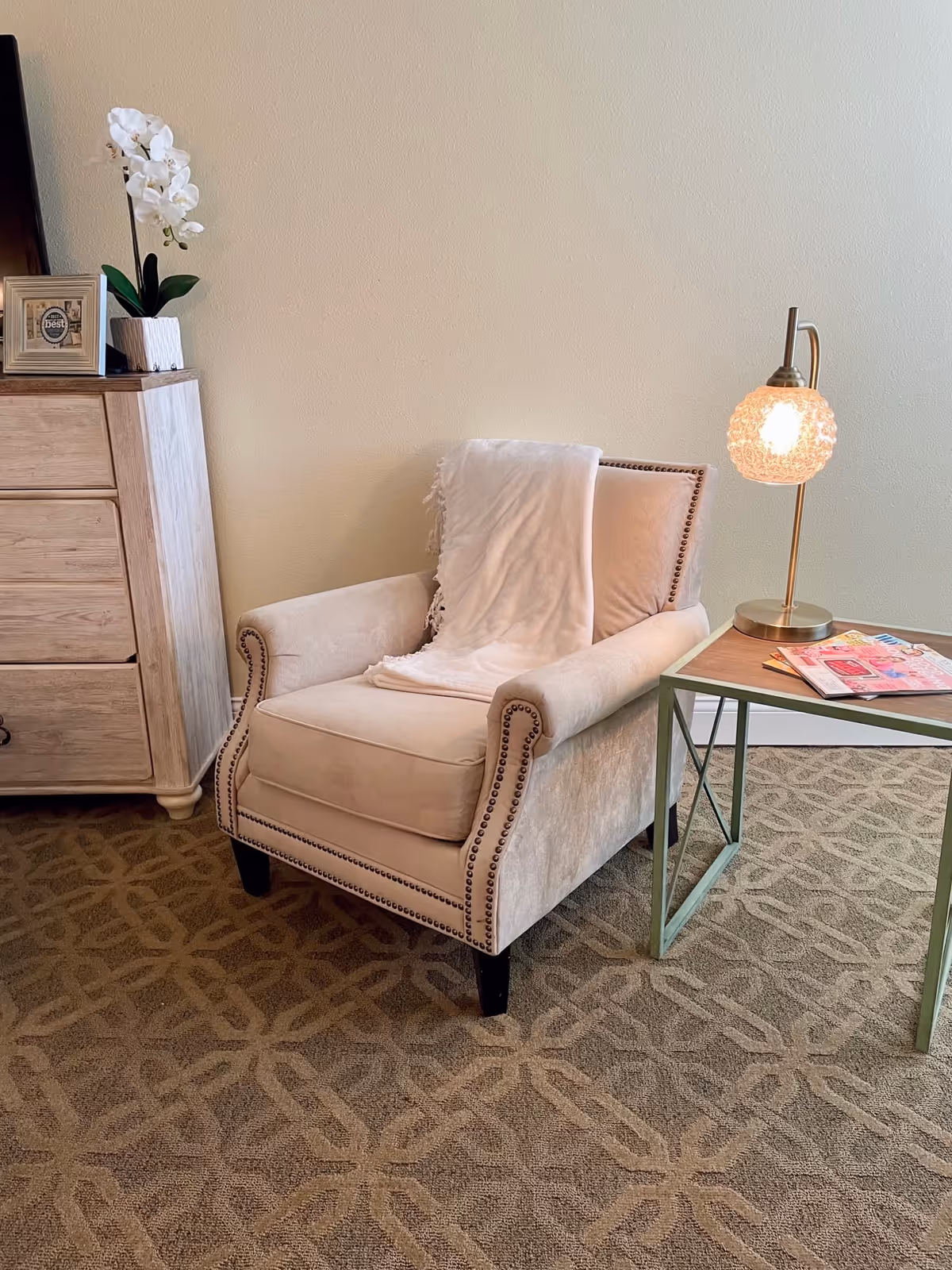 A cozy beige armchair with nailhead trim and a white throw blanket draped over the back, positioned next to a small side table with a decorative lamp and magazines on top. To the left, there is a light wood dresser with a white orchid plant and a framed picture on it. The floor is carpeted with a patterned design.