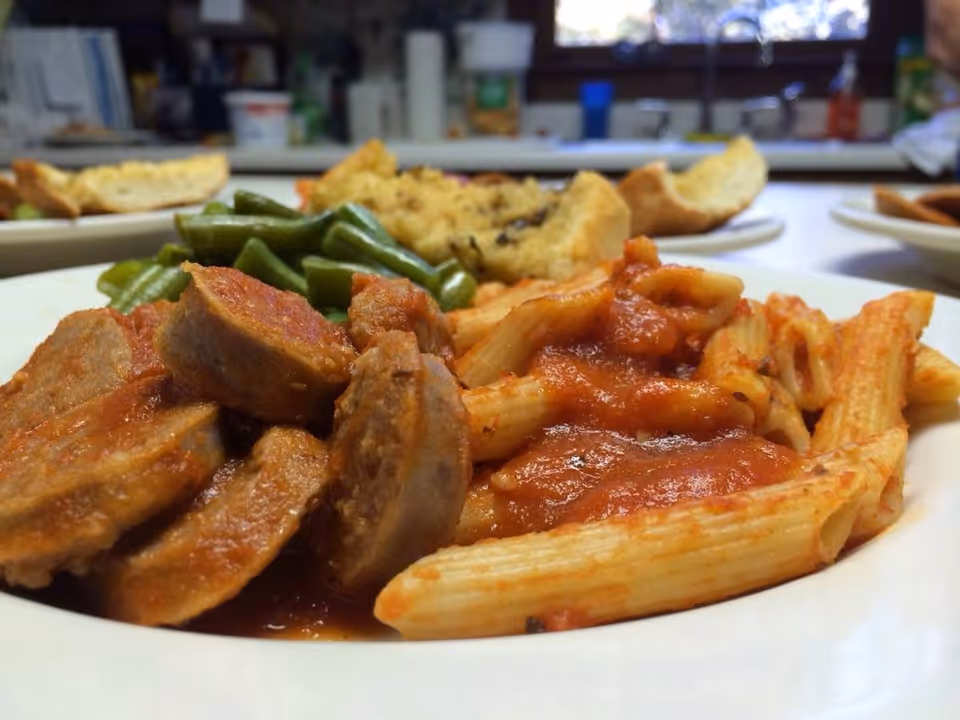 Close-up of a plate of food featuring penne pasta with tomato sauce, sliced sausage, green beans, and a piece of garlic bread in the background on a kitchen counter.